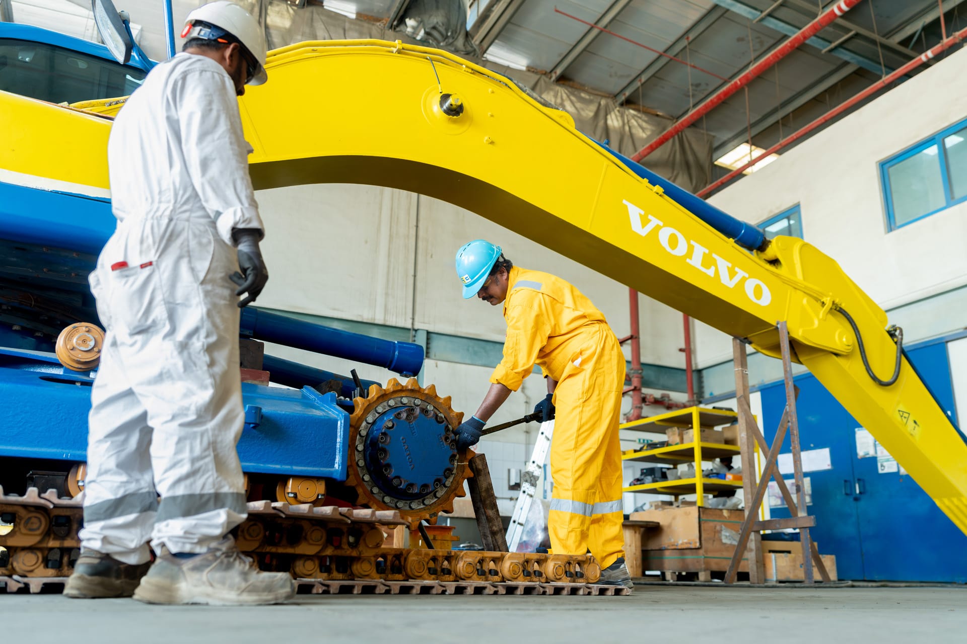 Low-angle view of a Volvo excavator boom sweeping over two workers servicing the machine's track and sprocket.
