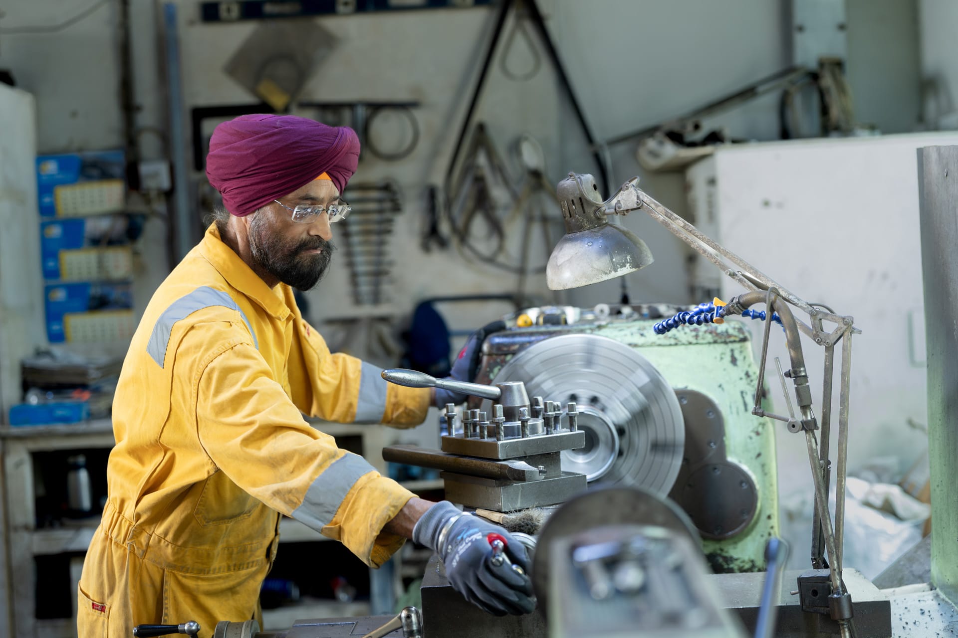 Machinist in a maroon turban and yellow coveralls operating a green manual metal lathe in a workshop.