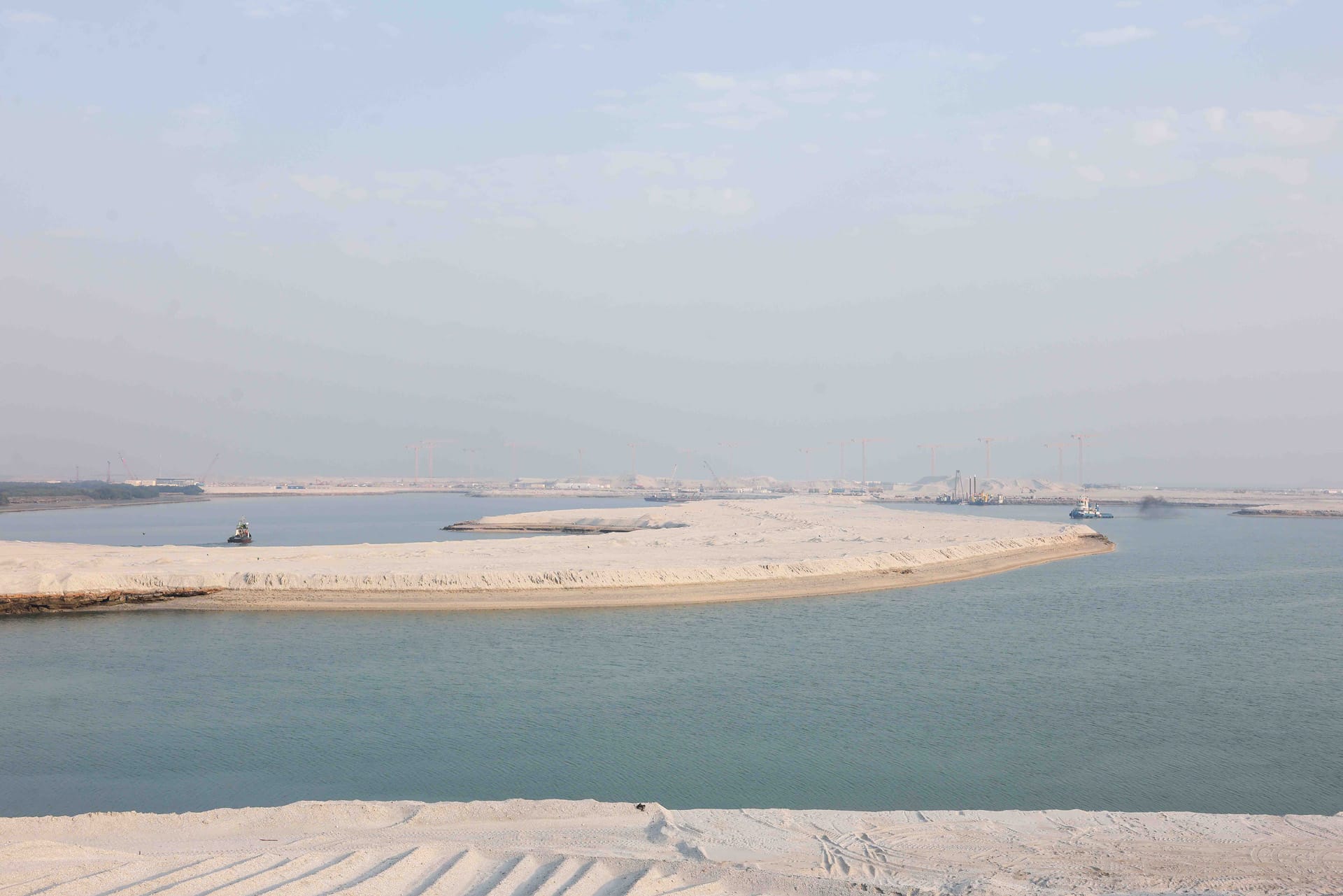Wide reclamation channel with tugs, a dredger and rows of tower cranes on the distant horizon.