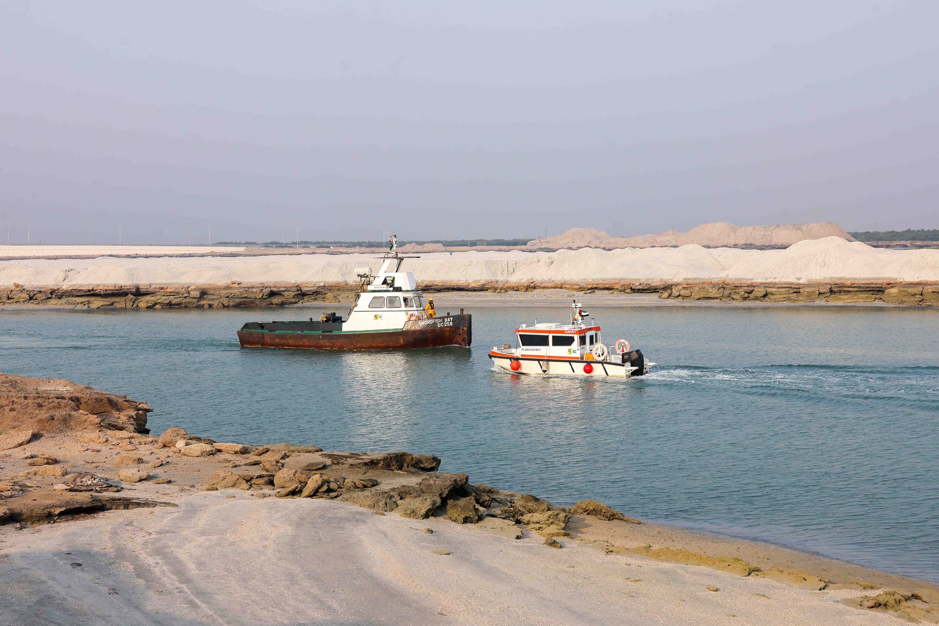 Two Gulf Cobla workboats moving through a dredged channel flanked by pale sand embankments.