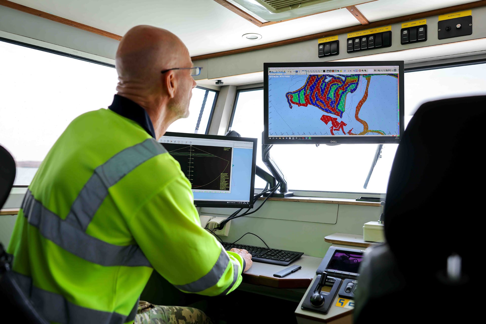 Operator studying dredge plan and depth profile screens at a wheelhouse workstation.