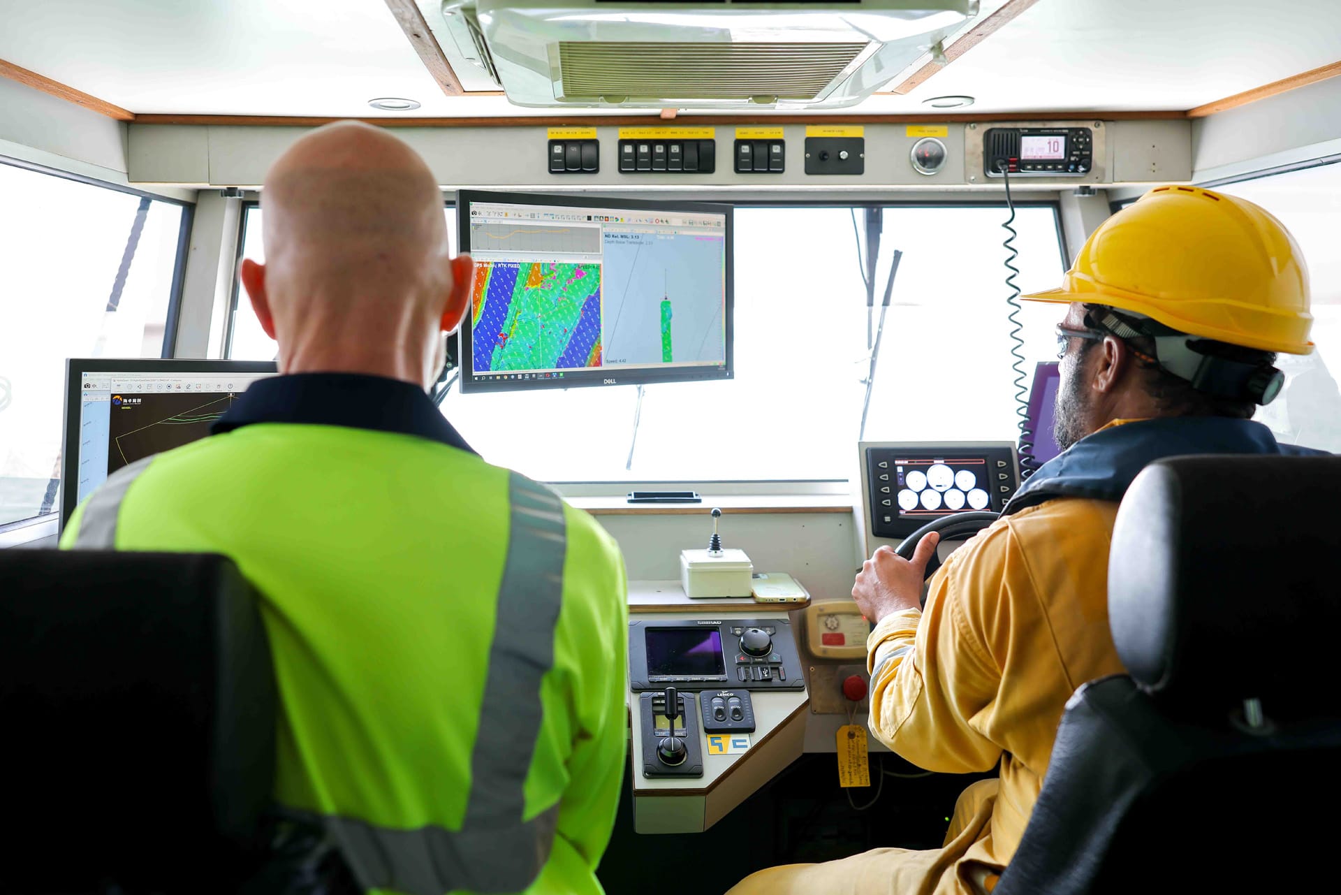 Two crew in a survey vessel wheelhouse, one at the survey monitors and one at the helm.