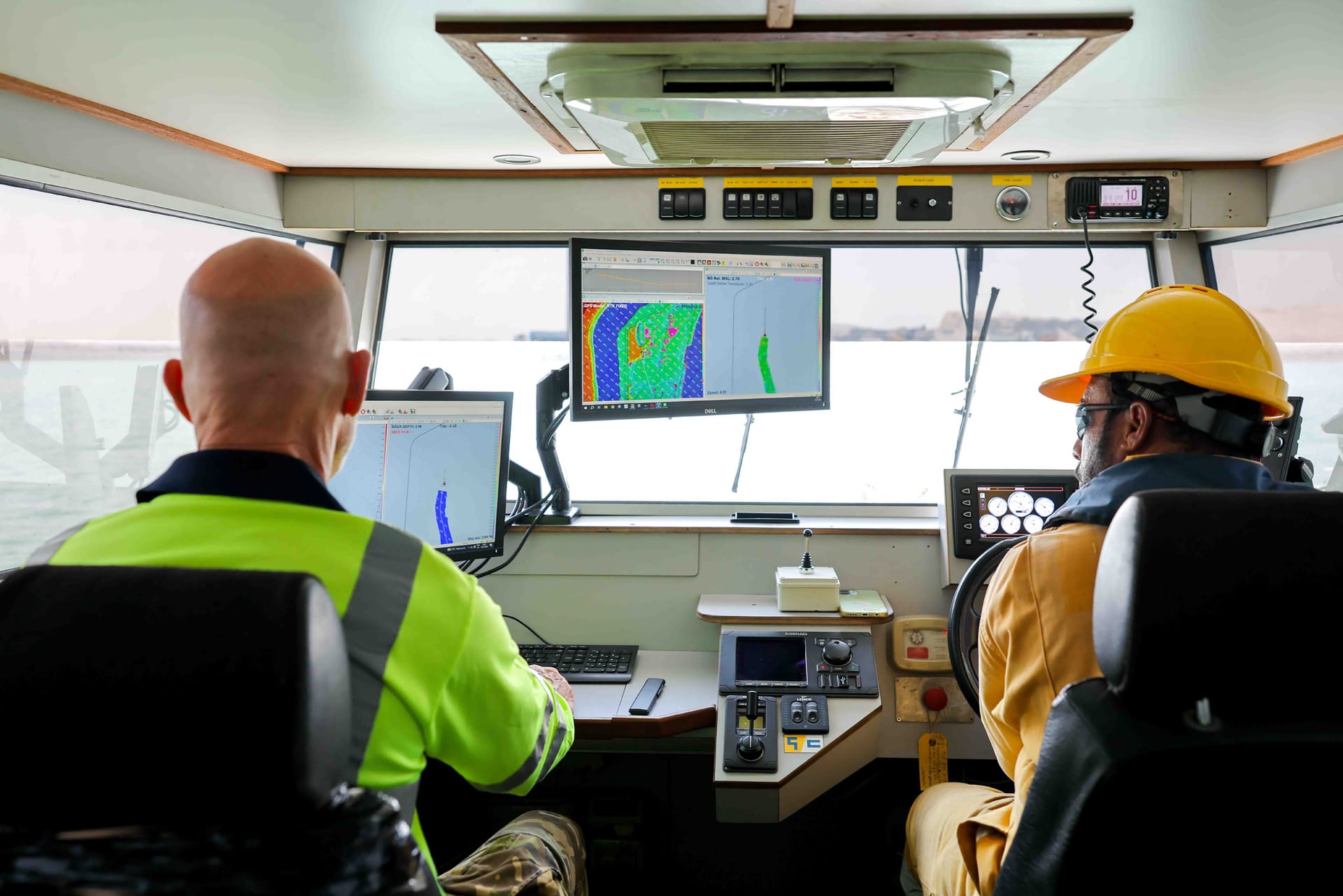 Survey vessel wheelhouse with a surveyor at the monitors and a helmsman in a yellow hard hat.