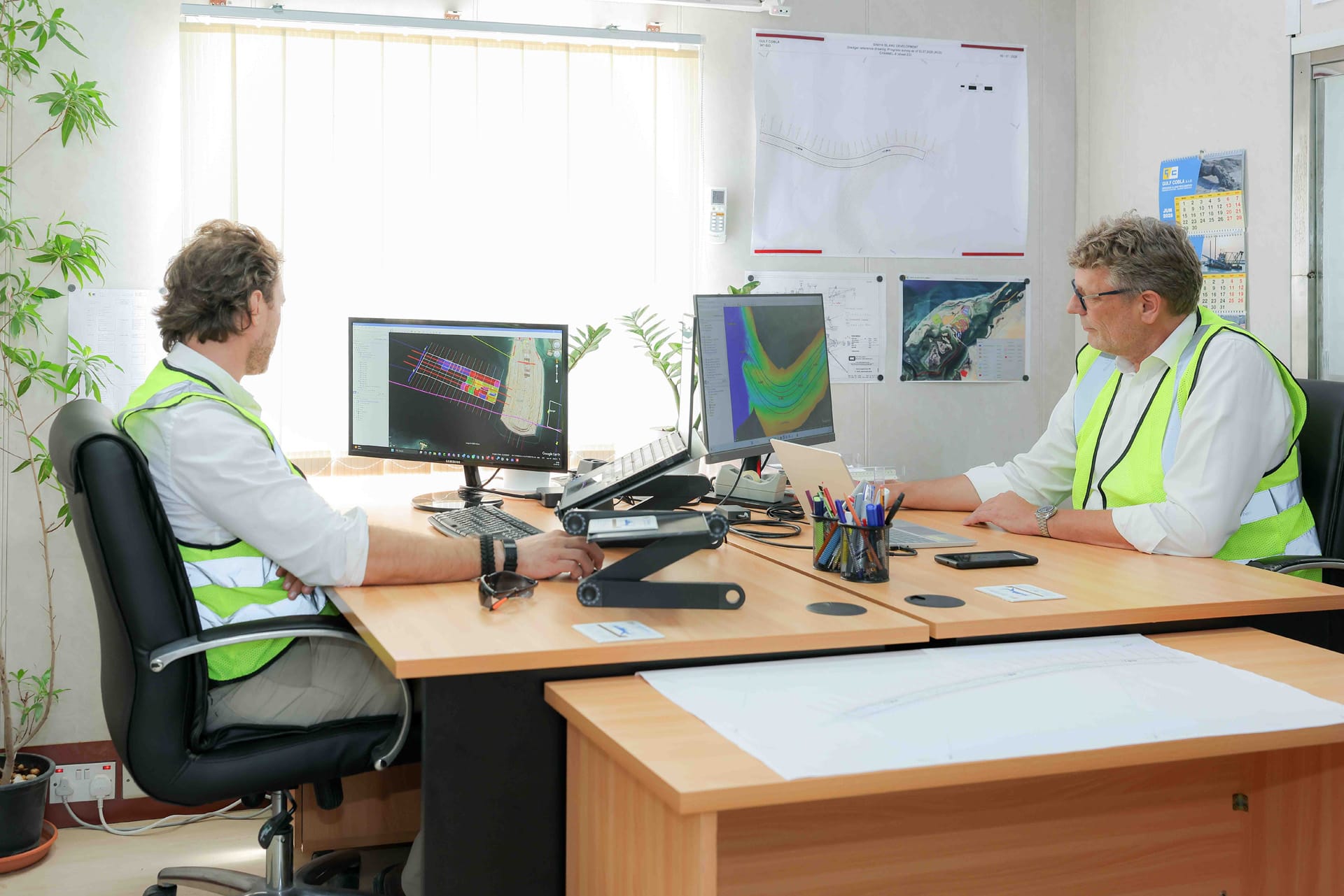 Two engineers at an office workstation reviewing dredge survey plots and bathymetry on dual monitors.