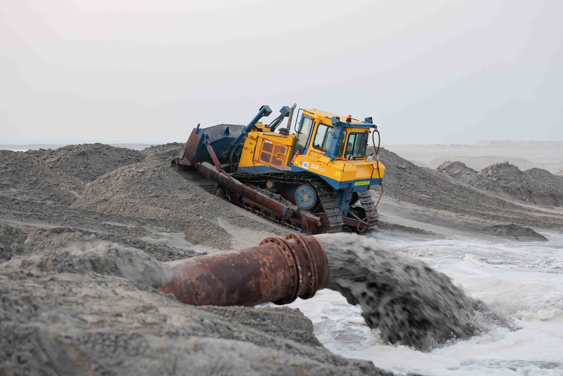 Gulf Cobla D6R XW dozer working on sand mounds beside a discharge pipe outfall at dusk.